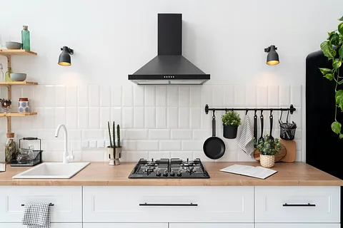 Moderne kjøkken med hvite fliser, sort ventilator, gasskomfyr og trebenk med planter og kjøkkenredskaper.